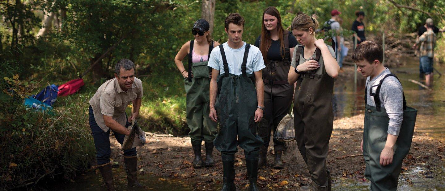 A group of students wearing overalls and boots, observing an instructor at the Biological Preserve.